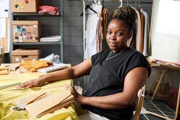 Young African American seamstress or fashion designer sitting by her workplace with textile and...