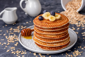 Oat milk pancakes. Stack of homemade pancakes served on a plate with banana slices, honey and fresh blueberry. Horizontal. Milk jug and oatmeal flakes on background.