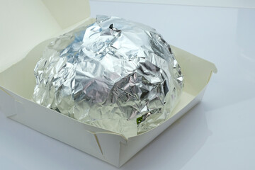 Hamburger in foil and cardboard box on a white background. The burger is wrapped in foil to keep it warm. Fast food delivery.