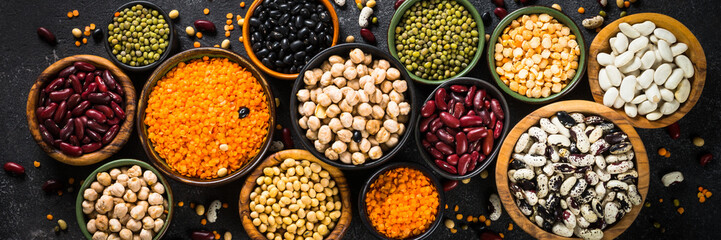Legumes, lentils, chikpea and beans assortment in different bowls on black stone table. Long banner...