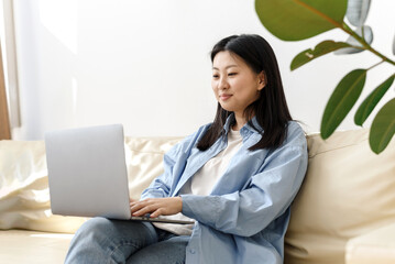 Smiling young asian female freelancer working from home, sitting on couch, using laptop, copy space. Relaxed korean young woman enjoying her weekend, surfing on internet on laptop