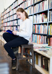 Intelligent little girl sitting on wooden stepladder reading interesting book in bookstore