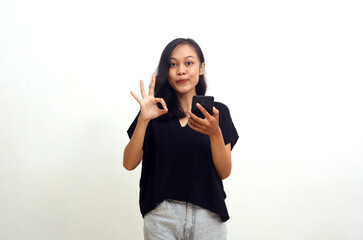 Young asian woman showing okay gesture while holding a cellular phone. Isolated on white background