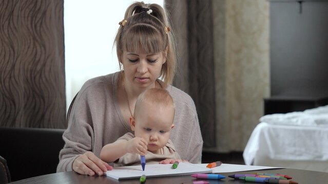 Mom, baby play together and draw at home in room. Young mother teaches cute baby son to draw with colored markers on paper sheet. Family adheres to early education sitting at table in bedroom at home.