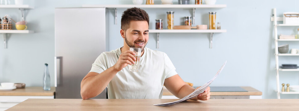 Handsome young man reading newspaper while drinking water in kitchen