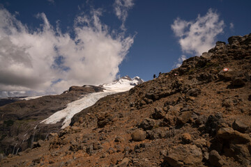 Alpine scenic. View of two hikers climbing up Tronador hill and glacier Castaño Overo in the Andes mountains in Patagonia Argentina. The rocky mountaintop and glacier ice field under a majestic sky.
