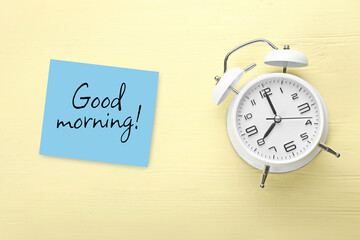 Alarm clock and paper with text GOOD MORNING on color wooden background, top view