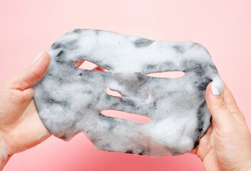 Female hands holding sheet of black bubble mask close-up top view, pink background.