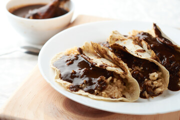 Tacos of mole with rice, placed on wood, in the background a piece of chicken