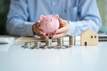 Businessman holding a piggy bank and a pile of coins in front with a calculator house to calculate...