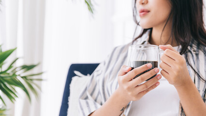 Crop image of woman's hands holding a cup of coffee in the living area with a green plant, Relaxing drinking a cup of hot coffee and enjoying a quiet time and indoor environment at cozy home.