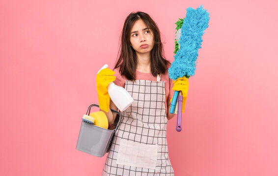 Boring young Asian woman wearing apron with yellow rubber gloves and holding bucket of cleaning supplies on pink background.