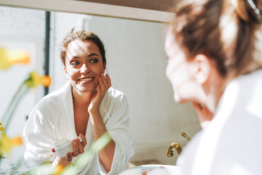 Beauty portrait of young smiling woman with dark long hair in white bathrobe put day nourishing moisturizer cream on clean fresh skin face and hands near mirror in bathroom at home