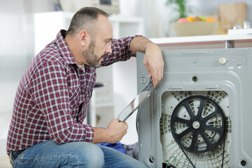 Fototapeta serviceman repairing washing machine looking at tablet obraz