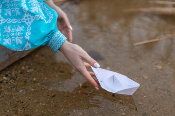 Beautiful young Ukrainian woman dressed in a blue embroidered dress lowers a paper boat. Close up of a girl lowers a boat into the water