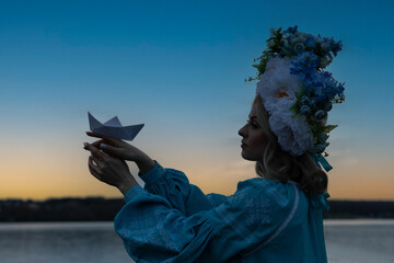 Young beautiful Ukrainian woman in a blue embroidered shirt, holding a paper boat made of white...