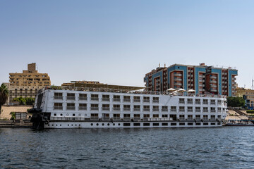 Naklejka premium The tourist ship is moored at the river bank. Cabin windows and railings are visible. On the upper deck there is an awning, umbrellas. Ripples on the water. City houses against the blue sky. Egypt