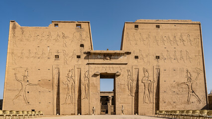 The Temple of Horus in Edfu. A high wall with huge carvings of gods against the blue sky. At the...