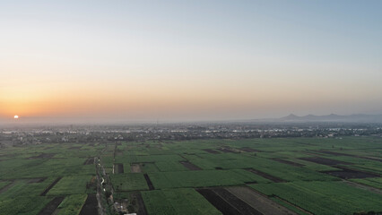 Aerial view of the Nile Valley at dawn. Below are green rectangles of cultivated fields. In the distance - a village, a river.  The sun rises in an orange-blue sky. Egypt. Luxor