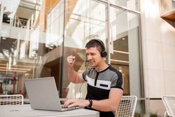 A young man or student or businessman is sitting at a table with a laptop and headphones. Rejoicing in the victory.