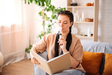 A beautiful young woman uses a disposable electronic cigarette on the couch at home reading a book....