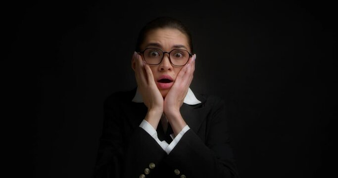 Frightened brunette in glasses and office clothes is shocked by what she saw. A business woman holds her palms to her cheeks in surprise, standing on an isolated black background