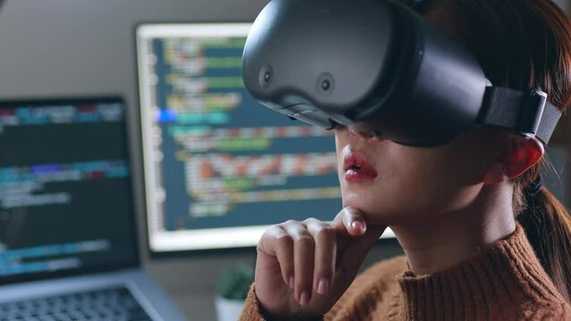 Young Asian woman, developer programmer, software engineer, IT support, wearing virtual reality headset developing and programming VR game application at night overtime.