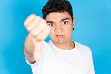 Sad negative hispanic teenager boy thumbs down looking at camera isolated on blue background.