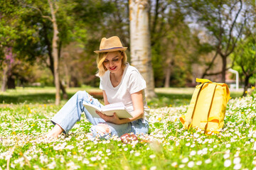 A young blonde girl in a hat reading a book in spring in a park in the city, vacations next to nature and next to daisies