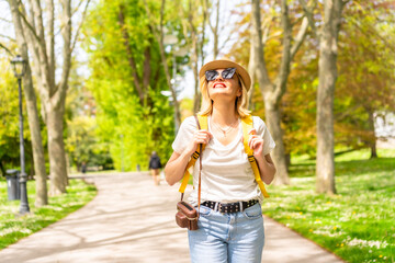 A tourist blonde woman with a hat and sunglasses walking in the spring in a park in the city, with the camera, enjoying the sun