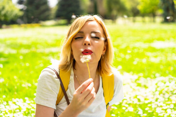 A smiling blonde woman blowing on the dandelion plant, wearing a hat and sunglasses sitting on the grass in spring next to daisies in a park in the city