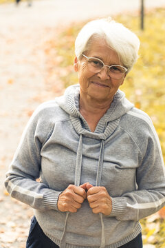 Elderly caucasian pensioner woman standing in a park, looking at camera, smiling, and touching her gray hoodie. Vertical outdoor shot. High quality photo