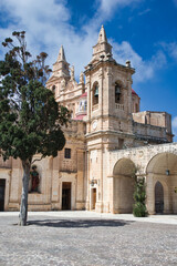 Fototapeta na wymiar Old church in Malta with big towers on a perfect blue sky background with a big tree in front casting its shadow over the yard creating a peaceful vacation scene