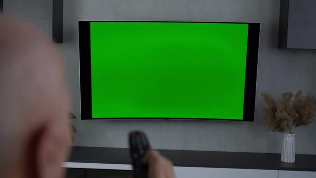 Elderly man changes channels on chroma key TV screen using remote control sitting on couch at home