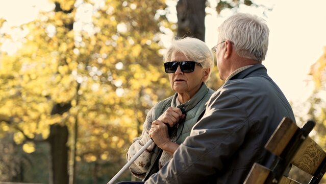 Senior retired caucasian couple sitting on the bench in the park, disabled wife in glasses and a stick park selective focus. High quality photo
