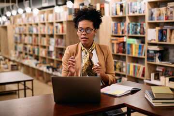 Black female teacher talks while holding online class over laptop from library.