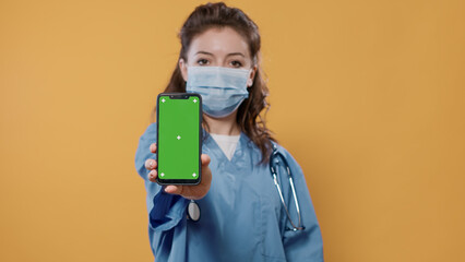 Portrait of medic in hospital uniform wearing surgical mask holding smartphone and texting showing green screen mockup at camera. Doctor with stethoscope and covid protection presenting copy space.