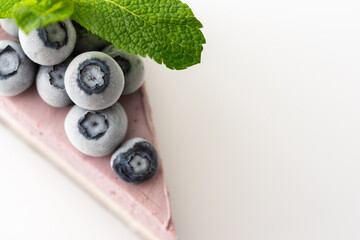 A piece of multi-colored delicious blueberry cheesecake decorated with mint and frozen blueberries close-up on a white background