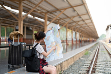 Fototapeta premium Young asian woman using generic local map, siting alone at train station platform with luggage. Summer holiday traveling or young tourist backpack traveler concept
