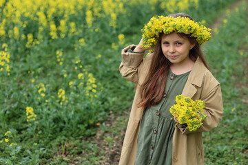 a cute six-year-old girl in a khaki cotton dress stands in a rapeseed field with a wreath of rapeseed flowers on her head