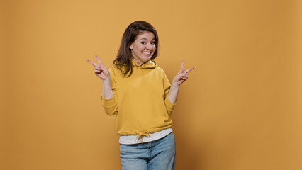 Portrait of casual smiling woman showing peace sign hand gesture in studio. Smiling girl showing victory sign celebrating on yellow background. Happy young adult making v sign with fingers.