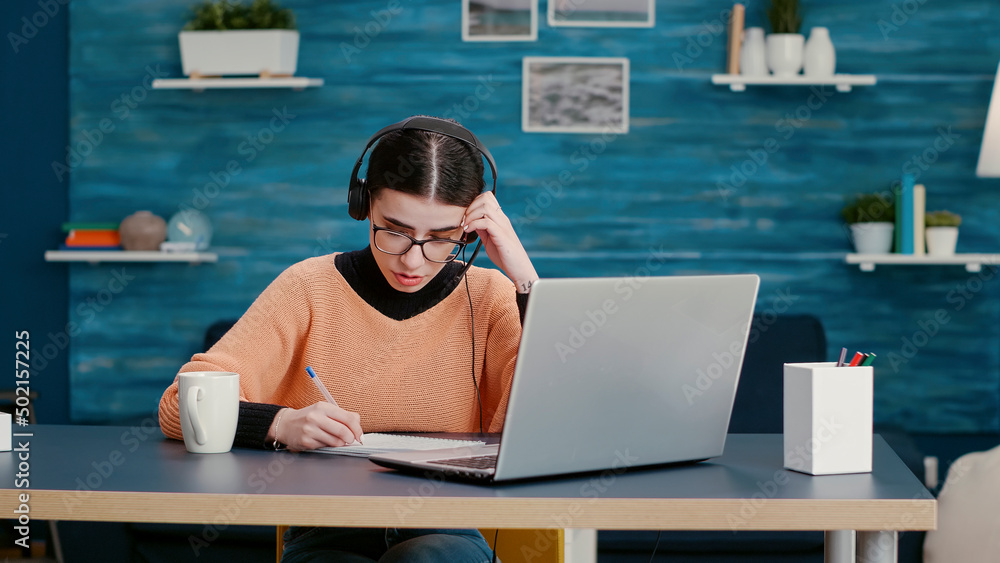 Poster woman with headphones talking on online video call and taking lecture notes on notebook, learning in