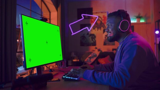 Excited Gamer Playing Online Video Game with a Mock Up Green Screen on His Powerful Personal Computer. Room and PC have Colorful Neon Led Lights. Cozy Evening at Home in Loft Apartment.