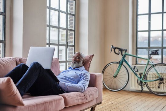Mature businessman using laptop lying on sofa at home