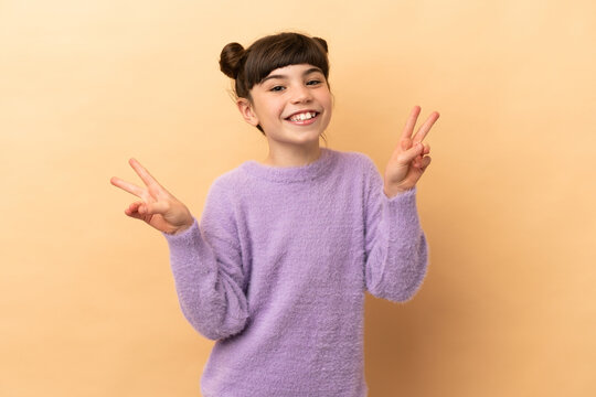 Little caucasian girl isolated on beige background showing victory sign with both hands
