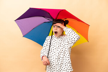 Little girl holding an umbrella isolated on beige background doing surprise gesture while looking to the side