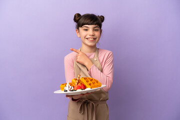 Little caucasian girl holding waffles isolated on purple background pointing to the side to present a product