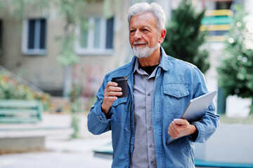 A casual senior businessman with coffee and a laptop in his hands walking outside.