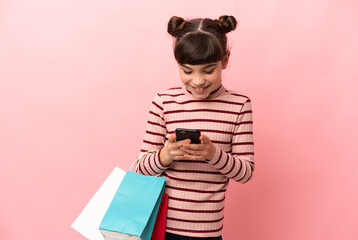 Little caucasian girl isolated on pink background holding shopping bags and writing a message with her cell phone to a friend
