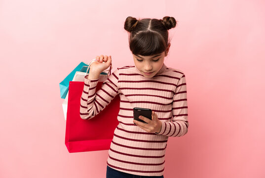 Little caucasian girl isolated on pink background holding shopping bags and writing a message with her cell phone to a friend